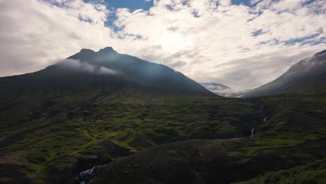 Rotierender-Zeitraffer-Von-Wolken-Und-Der-Sonne,-Die-Zwischen-Mehreren-Bergen-Durchbricht-Und-Lichtstrahlen-Und-Schatten-In-Island-In-4k-Wirft