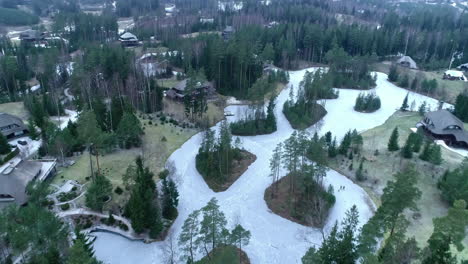 Aerial-flyover-frozen-lake-in-Amatciems-Park-with-forest-woodland-in-winter