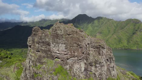 Hiker-on-Cliff-Edge-Mountain-Peak-overlooking-Kahana-Bay,-Oahu---Aerial-Drone-Flyover