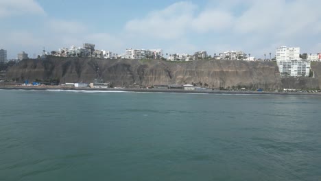 aerial-dolly-drone-shot-of-the-coast-in-miraflores-lima-in-peru-from-a-sea-with-calm-waves-towards-the-beach-with-various-buildings-and-rocks-in-the-background-on-a-sunny-day