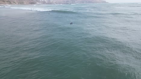 Aerial-panning-shot-of-the-beautiful-turquoise-sea-off-the-coast-of-miraflores-in-peru-lima-with-surfers-on-surfboards-in-the-water-overlooking-a-highway-along-the-rocks-in-the-background