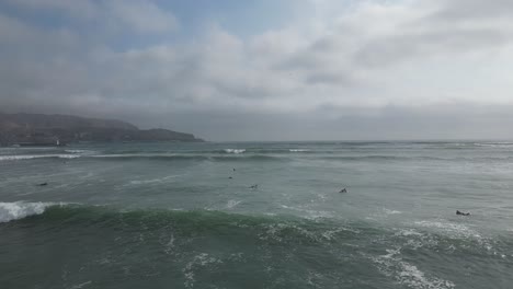 Wide-aerial-dolly-shot-of-the-calm-sea-in-miraflores-lima-in-peru-with-surfers-on-surfboards-in-the-water-and-a-view-of-the-cloudy-sky-with-mountains-in-the-background