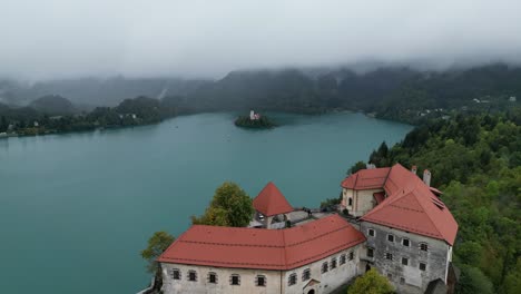 Drohne-Enthüllt-über-Bled-Castle-Niedrige-Wolken,-Die-Ferne-Berge-Bedecken