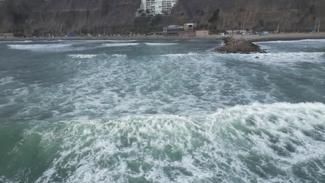 Slow-motion-aerial-dolly-shot-of-the-sea-with-waves-in-front-of-the-beach-of-miraflores-lima-in-peru-with-rocks-and-building-in-the-background
