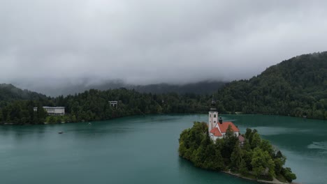 Lake-Bled-Slovenia-reverse-drone-aerial-view-misty-low-clouds-in-background