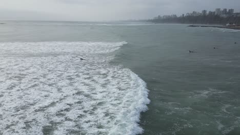 Aerial-panning-shot-of-the-sea-in-Lima-Peru-with-calm-waves-and-surfers-with-surfboards-in-the-water-on-a-cloudy-day