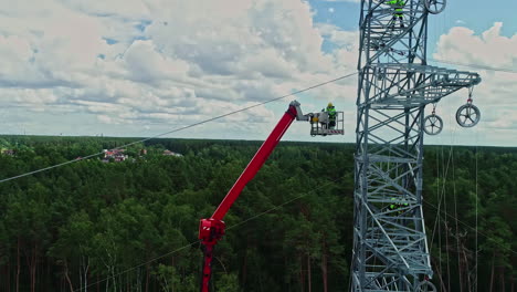 Static-aerial-drone-shot-of-one-among-the-three-technicians-is-coming-down-using-lifting-crane,-other-two-are-staying-up-while-working-on-high-voltage-electricity-tower-in-their-uniform