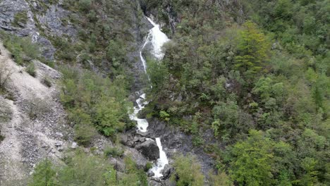 Waterfall-cascading-though-trees-Lake-Bohinj-Slovenia-rising-crane-drone-aerial-view