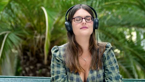A-young-woman-with-glasses-with-closed-eyes-listening-to-music-in-headphones-in-the-park-on-a-bench-against-the-backdrop-of-a-palm-tree