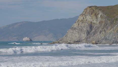 Stationary-shot-of-waves-crashing-into-the-side-of-a-cliff-with-mountains-on-the-background-located-in-Big-Sur-California-Beach