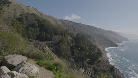 Panning-shot-from-mountain-side-to-the-Pacific-ocean-at-Big-Sur-California-beach-with-waves-crashing-on-a-rocky-shore