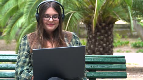Schöne-Studentin-Mit-Kopfhörern,-Die-An-Einem-Laptop-Auf-Einer-Bank-Im-Park-Arbeitet