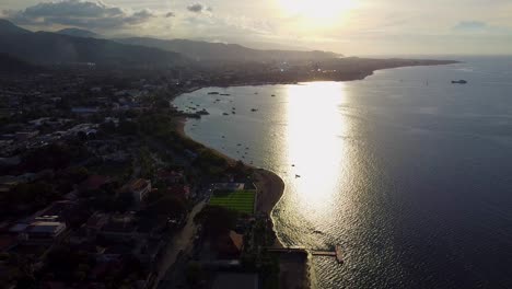 Aerial-drone-lowering-over-capital-city-Dili,-Timor-Leste-in-Southeast-Asia-with-beautiful-golden-sunlight-reflecting-off-the-ocean-water-and-boats-moored-in-calm-waters-along-waterfront