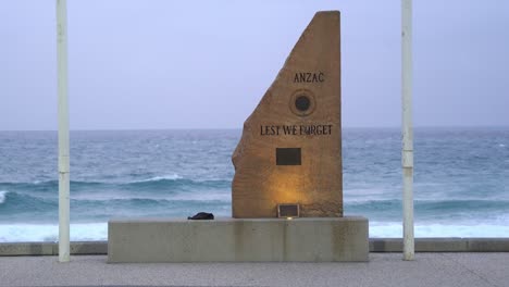 Close-up-to-ANZ-war-memorial-cenotaph-carved-with-Lest-We-Forget-with-rapid-and-vigorous-wave-crashing-on-to-the-shore-in-the-background-at-surfers-paradise,-Gold-Coast,-Queensland,-Australia