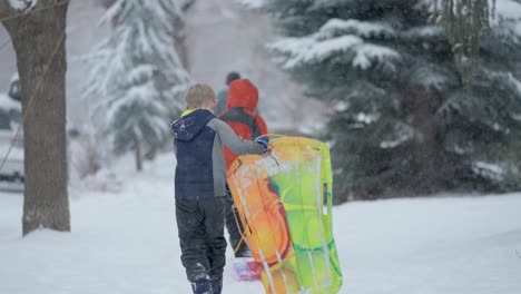 Young-white-male-walking-on-a-snow-covered-sidewalk-carrying-a-colorful-sled