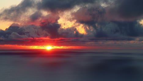 the-red-sun-sets-over-the-ocean-coloring-the-moving-clouds,-real-time-shot