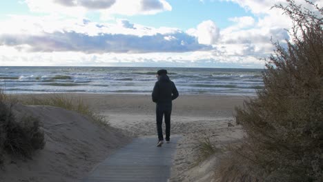 Back-view-of-caucasian-male-exploring-nordic-seaside-forest,-wooden-pathway,-man-walking-alone-towards-the-Baltic-sea-white-sand-beach,-sunny-day-with-clouds,-healthy-activity-concept,-wide-shot