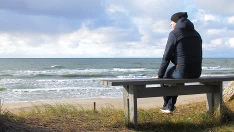 Back-view-of-caucasian-male-exploring-nordic-seaside-forest,-man-sitting-alone-on-the-gray-wooden-bench-on-the-beach,-coastal-pine-forest,-white-sand-beach,-healthy-activity-concept,-wide-shot