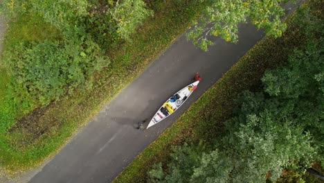 Drone-shot-of-Hikers-walking-with-canoe-across-a-straight-road-in-the-green-woods