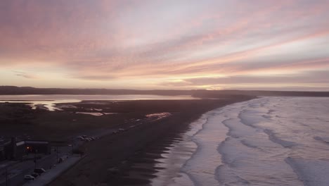 Dawn-sunrise-aerial-rises-above-beach-town-sand-dunes,-Tramore,-IRL