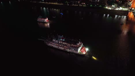 Aerial-view-overlooking-illuminated-steamboats-driving-on-the-Ohio-river,-nighttime-in-Cincinnati,-USA