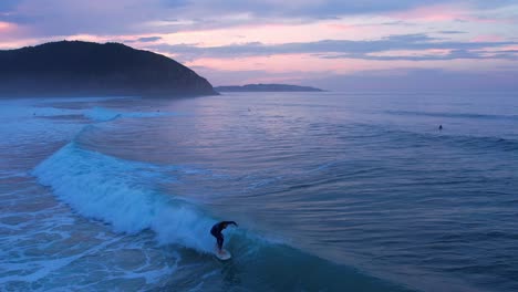 Drone-shot-of-a-surfer-riding-the-wave-at-Berria-beach-in-Cantabrian,-Spain-at-the-sunset