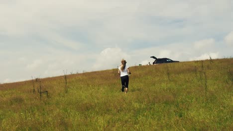 Long-haired-blonde-girl-running-up-a-hill-on-grass-in-slow-motion-with-clouds-in-the-background,-drone-follow-cam