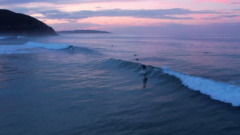 Drone-shot-of-a-surfer-of-a-surfer-catching-a-wave-at-Berria-beach-in-Cantabrian,-Spain-at-the-sunset