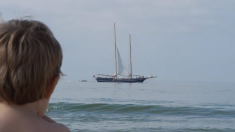 Niño-Caucásico-Mirando-Un-Barco-Vintage-Navegando-En-La-Superficie-Del-Mar-Desde-La-Distancia