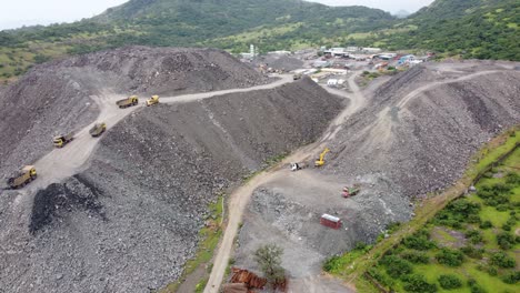 Aerial-view-of-a-mountain-of-debris-or-a-rubble-heap-with-dumper-trucks-unloading-more-rock-debris-from-a-tunnelling-project-in-a-hilly-region-impacting-the-local-ecology-and-terrain