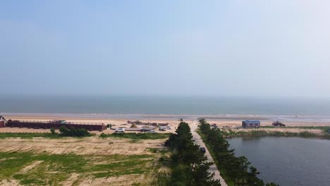 Aerial-ascending-pedestal-view-of-Nanhai-sandy-beach-with-pond-on-the-right-side-of-the-road-and-grass-of-the-left