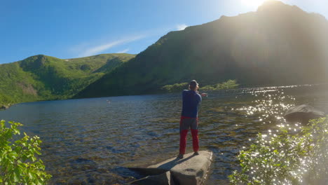 Man-using-fishing-rod-in-a-mountain-lake-on-a-nice-sunny-day,-static-shot-with-lens-flare