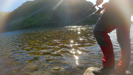 Low-static-shot-of-a-person-casting-a-fishing-line-into-a-river-then-reeling-it-in,-with-a-mountain-in-the-background-and-sun-flare-with-light-sparkling-off-the-water
