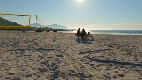 Dos-Turistas-Disfrutando-De-La-Vista-En-La-Playa-De-Flakstad,-Almorzando-Junto-A-La-Red-De-Voleibol-De-Playa