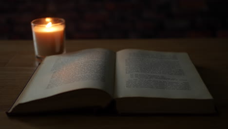 Ein-Altes-Buch-Auf-Einem-Tisch,-Beleuchtet-Von-Einer-Flackernden-Kerze-In-Einem-Dunklen-Raum