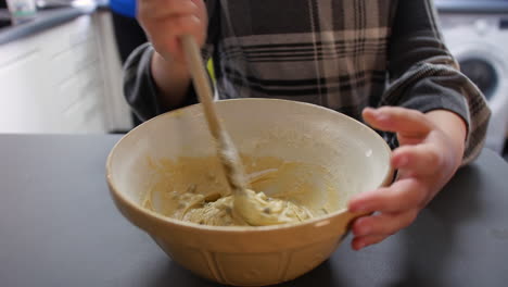 A-girl-mixing-cookie-dough-cake-mixture-in-a-bowl-at-home-in-a-kitchen-with-a-wooden-spoon