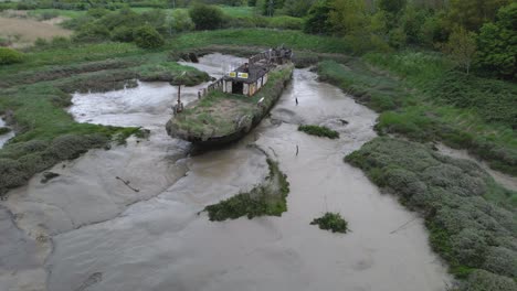 Overgrown-abandoned-boat-wreck-stranded-in-muddy-Wat-Tyler-riverbed-aerial-pull-away-view