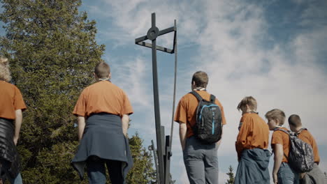 Young-scouts-clothed-in-orange-shirts,-some-wearing-backpack,-standing-in-front-of-a-statue-and-a-big-spruce-tree