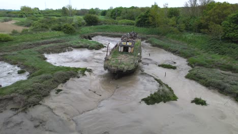 Abandoned-overgrown-boat-wreck-stranded-in-Wat-Tyler-muddy-riverbed-aerial-orbit-view