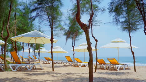Natural-sand-tropical-beach-and-sea-wave-with-yellow-beach-bench-under-white-parasol-umbrella-and-trees-on-tropical-island-beach