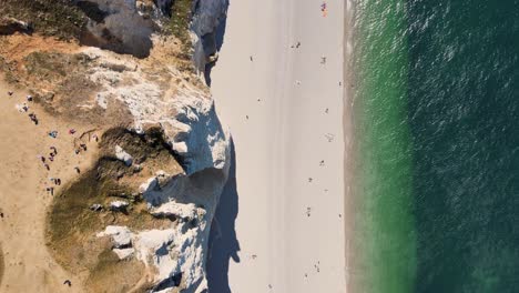 Aerial-top-down-flight-over-sandy-beach-along-cliff-coastline-during-sunny-day---People-walking-along-shore-of-sea---Etretat,-France