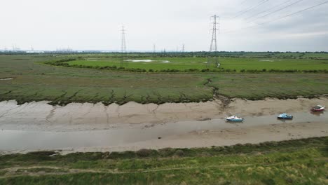 Flying-over-muddy-riverbank-marshland-towards-electric-supply-pylon-towers,-Basildon