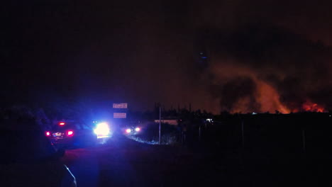 Police-vehicles-patrol-near-forest-fire-at-night,-red-flames,-black-columns-of-smoke-rise