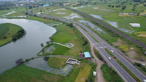 Cinematic-aerial-drone-shot-of-vast-farming-land-flooded-in-water-beside-passing-cars-and-heavy-vehicles-on-the-four-lanes-highway-and-a-railway-track-beside-the-highway-surrounded-by-mountains-at-far