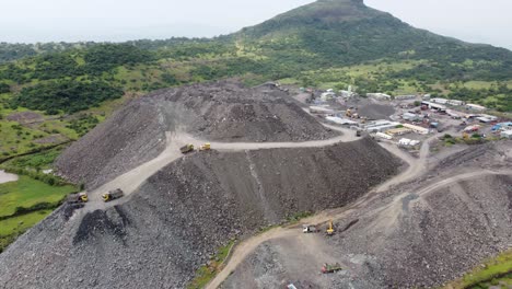 Aerial-drone-shot-flying-away-from-mountain-of-loose-debris-with-dumper-trucks-unloading-more-rock-waste-from-a-tunnel-project-in-a-hilly-region-impacting-the-local-ecology-and-terrain