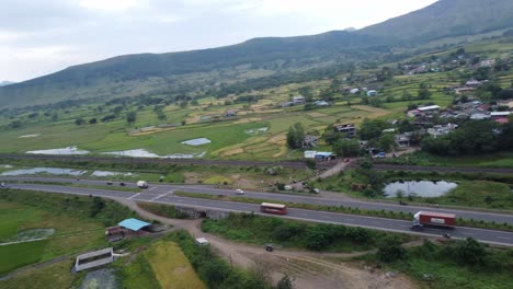 Cinematic-aerial-drone-shot-circling-around-passing-cars-and-heavy-transportation-vehicles-on-the-four-lanes-Indian-highway-and-railway-track-beside-it-surrounded-by-small-village-and-mountains