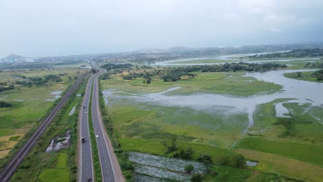 Cinematic-aerial-drone-shot-of-vast-farming-land-flooded-in-water-beside-passing-cars-and-heavy-vehicles-on-the-four-lanes-highway-and-a-railway-track