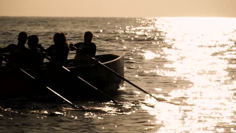 Silhouette-Motivierter-Menschen,-Die-Bei-Sonnenuntergang-Oder-Sonnenaufgang-Gemeinsam-Auf-Einem-Boot-Rudern