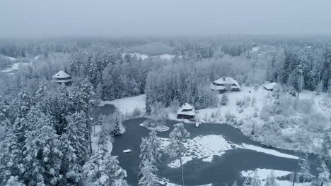 Drone-flying-over-winter-landscape-with-trees-and-homes-covered-in-fresh-snow-and-frozen-lake