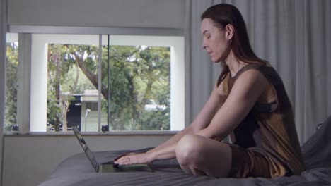 a-female-model-is-sitting-on-bed-and-working-on-her-laptop-computer,-slow-motion-close-up